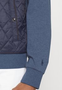 Marineblauwe doorgestikte buitenlaag met een gladde blauwe pullover-mouw, voorzien van een minimalistisch geborduurd logo vlak bij de manchet en een ritssluitingzak.