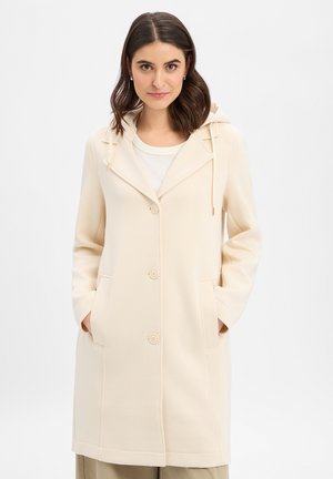 Kobieta o ciemnych włosach, ubrana w beżowy płaszcz z kapturem, z guzikami i kieszeniami, stojąca z rękami w kieszeniach na jednolitym tle.