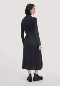 Langes, dunkelgraues Strickkleid mit einem hohen Kragen, langen Ärmeln und Seitenschlitzen, kombiniert mit schwarzen Stiefeletten. Glatte Textur.