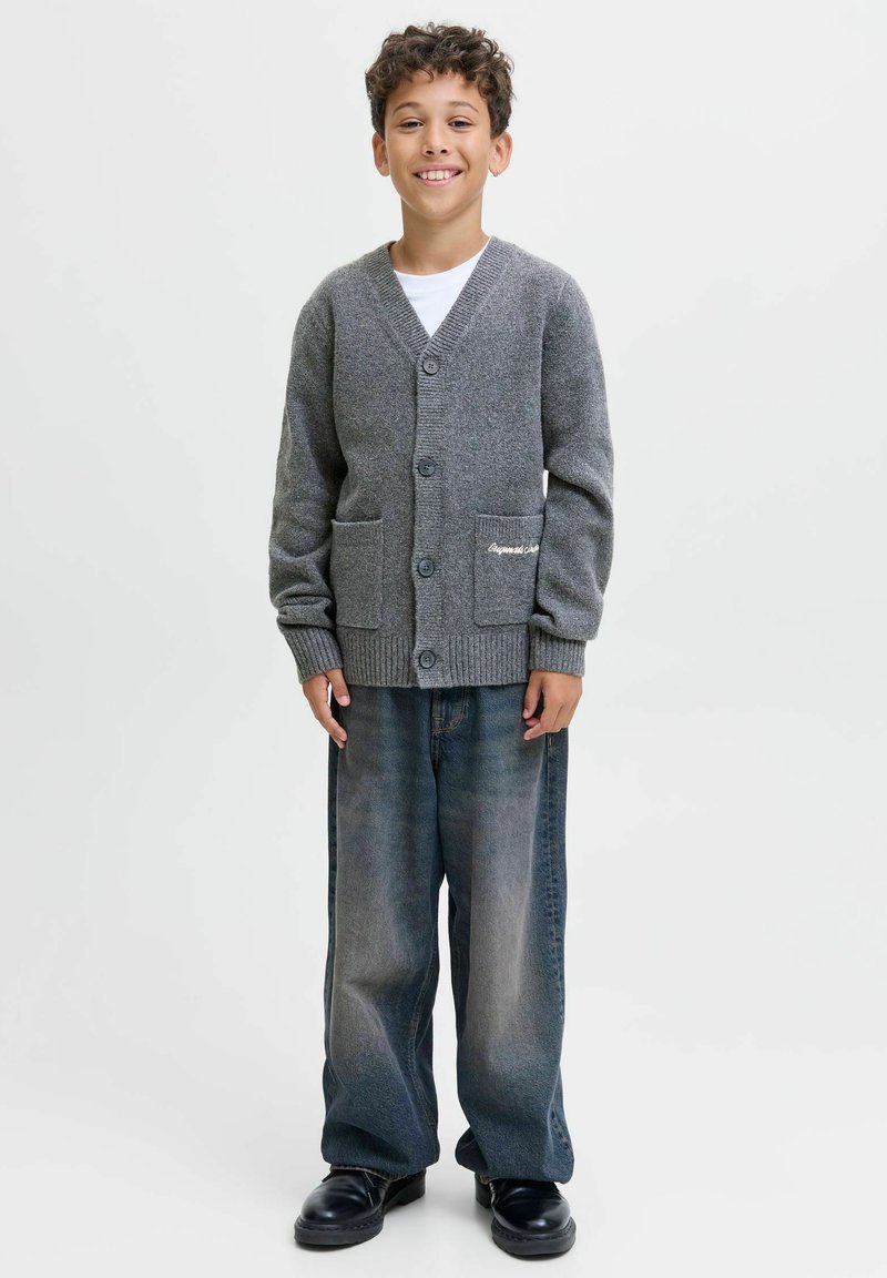 Pelēks adīts kardigans ar V-veida dekoltē, priekšējām kabatām un pogām. Apvienots ar platām, izbalējušām zilu džinsu biksēm un melniem apaviem.