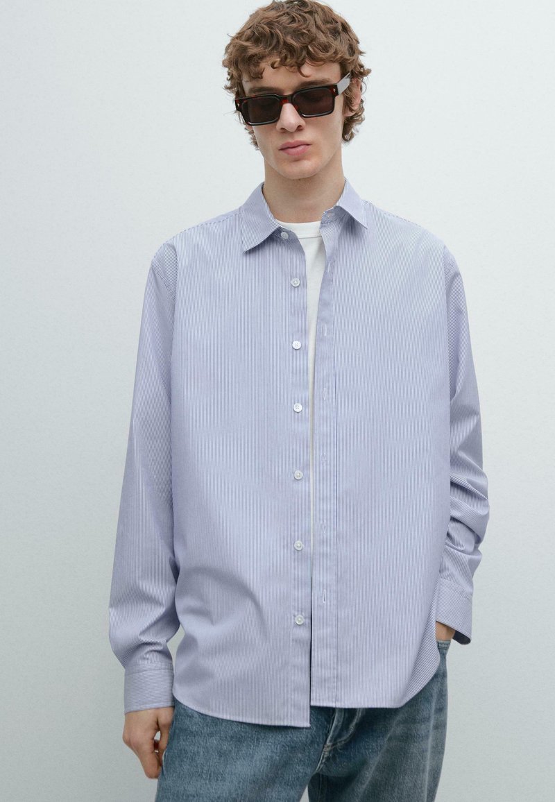 Camicia a righe azzurro chiaro con maniche lunghe e colletto abbottonato, indossata sopra una T-shirt bianca, abbinata a jeans di denim scuro.