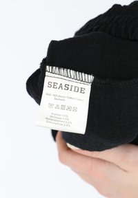 Schwarzes Baumwollkleidungsstück mit einer Textur, die leichte Faltenbildung zeigt. Ein Pflegeetikett mit der Aufschrift "SEASIDE" enthält Pflegehinweise und Anweisungen in mehreren Sprachen.