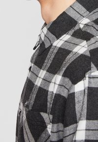 Gros plan d'une personne portant une chemise à carreaux noire, blanche et grise, boutonnée, avec un col et une poche poitrine.