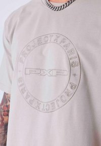 Camiseta de manga corta color beige claro con un logo circular en relieve que presenta "PROJECT PARIS" y "PXP" grabados en el centro. Textura de tela suave.