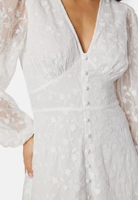 Vit broderad klänning med genomskinliga långärmar, med blommönster, V-ringning och knapprad framtill.