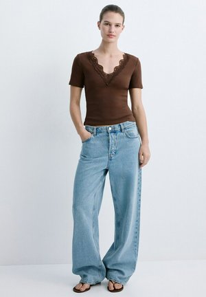 Tânără stă în fața unui perete simplu, purtând un top maro cu mâneci scurte, cu decolteu în V din dantelă și blugi largi, bleu deschis, având mâinile în buzunare.