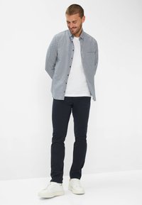 Hemd mit einem blau-weißen Karomuster, kombiniert mit navyfarbenen Hosen und weißen Sneakers. Das Model steht mit den Händen hinter dem Rücken.