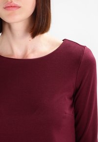 Bordeaux långärmad topp, mjukt material, rund halsringning, figurnära stil, subtil sömdekoration nära axeln, minimalistisk design.