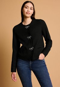 Čierna textúrovaná bunda s okrúhlym výstrihom, tromi dekoratívnymi striebornými mašľami a mierne rozstrapkaným lemom. V kombinácii s modrými džínami.