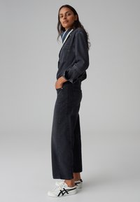 Giacca in denim scuro con tasca, abbinata a jeans larghi scuri. Sneakers bianche con accentuazioni nere. Design semplice e casual.