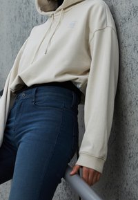 Felpa beige con cappuccio e logo, abbinata a jeans a vita alta blu scuro. Il tessuto sembra morbido, con una texture liscia.