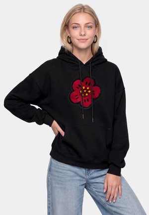 Giovane donna con capelli biondi indossa una felpa nera con cappuccio con un grande motivo floreale rosso, abbinata a jeans azzurri chiari.
