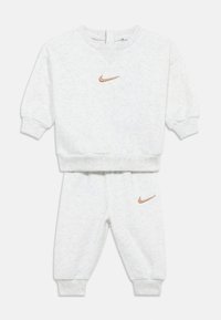 Šviesiai pilka sportinė apranga iš megztos audinio medžiagos. Komplekte yra suspaustos rankogaliai ir mažas oranžinis Nike logotipas ant krūtinės ir kairiojo kelnių šlaunies.