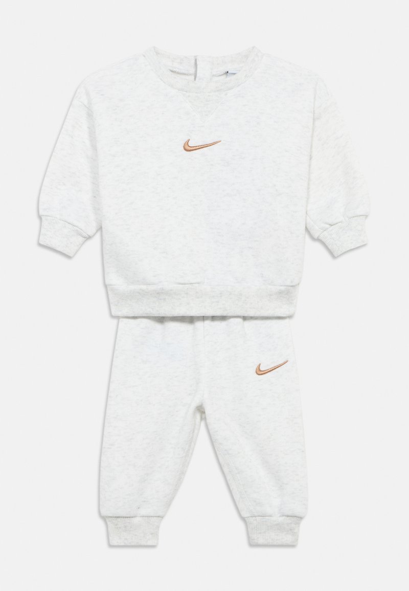 Šviesiai pilka sportinė apranga iš megztos audinio medžiagos. Komplekte yra suspaustos rankogaliai ir mažas oranžinis Nike logotipas ant krūtinės ir kairiojo kelnių šlaunies.