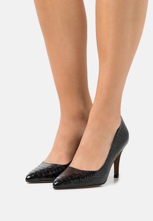 Femme portant des chaussures à talons hauts noires texturées avec des pointes, montrée de mi-mollet jusqu'aux pieds sur un fond blanc.