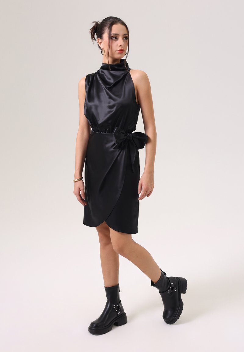 Robe en satin noir avec un décolleté drapé, une taille nouée en accent, et un design enroulé sur le côté, associée à des bottines noires avec des boucles.