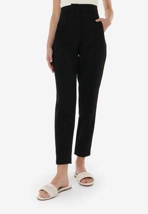 Black high-waisted trousers with a slim fit, featuring front pockets and subtle pleats. Paired with light-colored flat sandals.