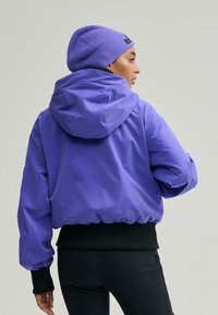 Lila vízálló dzseki kapucnival, rugalmas szegéllyel és kontrasztos fekete mandzsettákkal. Egy hozzáillő, bordázott lila sapkával viselve.