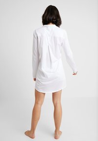 Abito bianco a maniche lunghe con colletto, dotato di una piega posteriore e orlo curvo. Realizzato in tessuto leggero con una texture liscia.