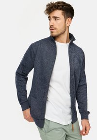Marine blå zip-up jakke i tekstureret stof, med en høj krave, sidelommer og et logo på brystet. Båret over en hvid t-shirt.
