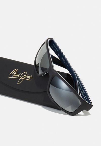 Occhiali da sole neri Maui Jim con lenti grigie oversize, finitura opaca e dettagli in argento, esposti su una custodia nera con logo.