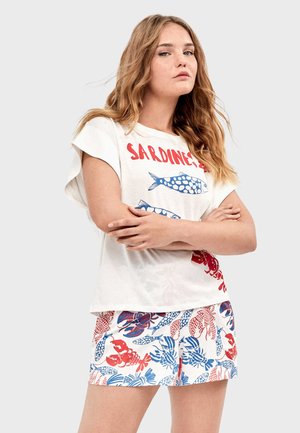 Jonge vrouw met lang haar die een wit bovenstuk draagt met rood- en blauwvisprint, en bijpassende korte broek met patronen van kreeften en vissen.