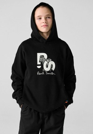 Junge Person mit schwarzem Hoodie mit PS-Logo und Zebragrafik, Hände in den Taschen, schaut leicht seitlich vor einfachem Hintergrund.