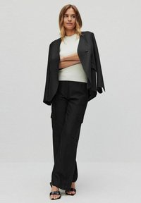 Zwarte blazer met een losse pasvorm, gelaagd over een witte geribbelde top en zwarte wide-leg cargobroek. Sandalen hebben een gespaccent.