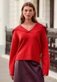 Red knitted sweater with a wide V-shaped neckline and dropped shoulders, paired with a dark leather skirt. The sleeves are ribbed at the cuffs.
