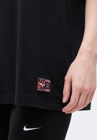 Το χέρι ακουμπά κοντά στη μαύρη ρίγα του μπλουζιού Nike με μικρό πολύχρωμο λογότυπο. Μαύροι κολάν με λευκό λογότυπο Nike ορατό από κάτω.