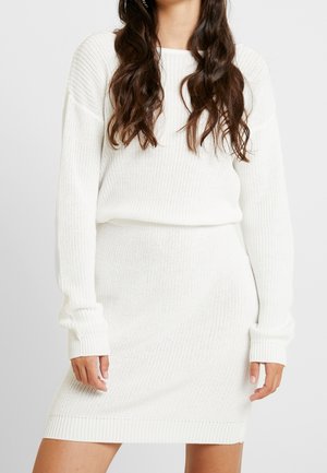 Mujer que lleva un suéter de punto blanco de manga larga y una falda de punto a juego, con ondas sueltas en su cabello.