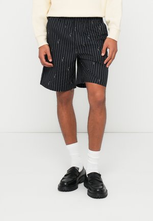 Pantaloni corti a righe nere con vita elastica, caratterizzati da un motivo sottile. Indossati con calze bianche e mocassini neri. Tessuto testurizzato.