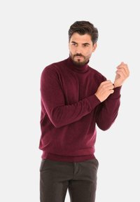 Maglione a collo alto bordeaux realizzato in tessuto morbido, dotato di maniche lunghe e polsini a coste. Indossato con pantaloni scuri.