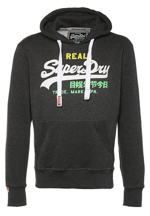 Mężczyzna ubraný w ciemnoszarą bluzę z kapturem Superdry, jasnoszare podarte dżinsy, czarne wysokie trampki i niebieski plecak, idący z jedną ręką w kieszeni.