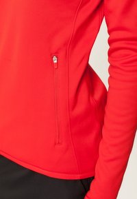 Chaqueta roja con cremallera fabricada en tejido suave, con un bolsillo lateral con cremallera, diseño ajustado y mangas largas.