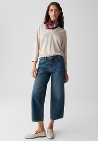 Beige Langarmoberteil, hoch-taillierte blaue Jeans und cremfarbene Loafers. Burgunderfarbener gemusterter Schal um den Hals gebunden. Einfarbiger Hintergrund.