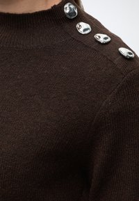 Gros plan sur l'épaule d'un pull marron en maille avec quatre boutons argentés brillants disposés en rangée près du col.