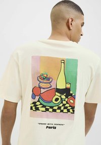 Cremefarvet t-shirt med et farverigt grafisk motiv af et bordopdækning med frugter og drikkevarer; baggrund med ternet mønster. Tekst under designet.