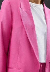 Růžový upravený blazer vyrobený z texturované tkaniny, s ostrými klopami a jednou kapsou, v kombinaci s bílým tričkem s kulatým výstřihem pod ním.
