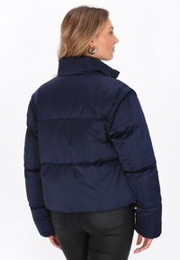 Tengerészkék puffos dzseki állógallérral, steppelt dizájnnal és cropped fazonnal. Fényes textúrával és cipzáras oldalzsebekkel rendelkezik.