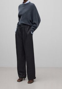 Grijze gebreide trui met verlengde schouders, gecombineerd met hooggetailleerde zwarte wijde broekspijpen. Model draagt zwarte sandalen met hakken en bandjes.