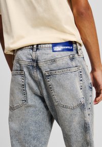 Jean en denim bleu délavé avec une coupe droite, comportant deux poches arrière et un patch logo bleu au niveau de la taille.