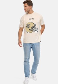 Camiseta color crema con un gráfico vintage del casco de los New Orleans Saints, de mangas cortas y cuello redondo, combinada con jeans azul claro.