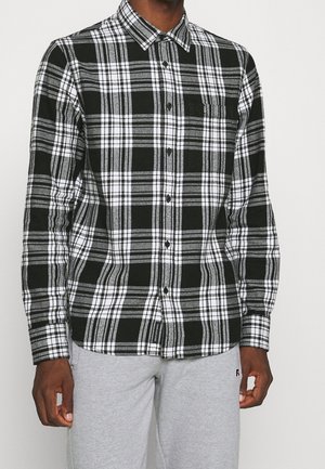 Homme portant une chemise à carreaux noire, blanche et grise à boutons avec manches longues et un pantalon de survêtement gris clair, debout devant un fond uni.