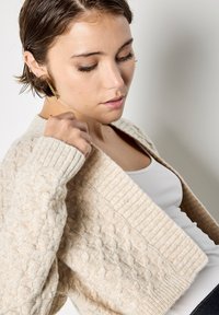 Cárdigan beige de punto con un patrón texturizado y detalles acanalados, con cuello chal y parte delantera abierta, llevado sobre una camiseta blanca.