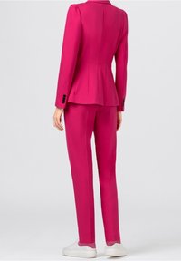 Fuchsiový sako na míru s peplum pasem a manžetami na dva knoflíky, ve spojení s ladícími kalhotami a bílými teniskami. Hladká struktura látky.