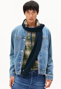 Veste en denim avec des boutons en métal, portée sur une chemise à carreaux verte et marron. Comprend une écharpe en tricot foncé enroulée autour du cou.