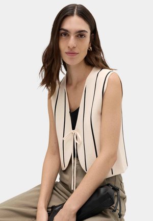 Donna con capelli castani alla lunghezza delle spalle indossa un gilet crema senza maniche con strisce verticali nere sopra una maglia nera, tenendo in mano una pochette nera.