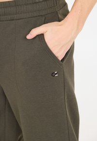 Olive green sweatpants featuring a ribbed waistband, side pockets, and a small embroidered logo on the left hip. Soft cotton material.