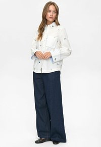 Vrouw in een witte blouse met kleine kleurrijke geborduurde details, donkere wijde spijkerbroek en zwarte gestructureerde instappers.